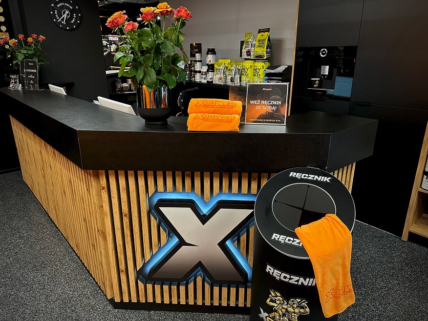 Recepcja klubu fitness z widocznymi ręcznikami do wypożyczenia oraz barem w tle.