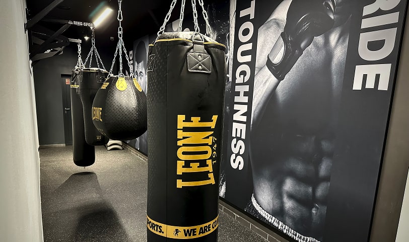 Strefa boksu z workami treningowymi oraz grafiką na ścianie przedstawiającą bokserów i motywujące hasła.