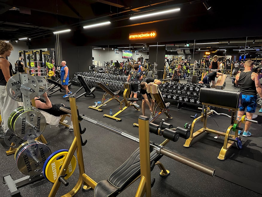 Strefa ciężarów z osobami ćwiczącymi ze sztangami i hantlami na siłowni.