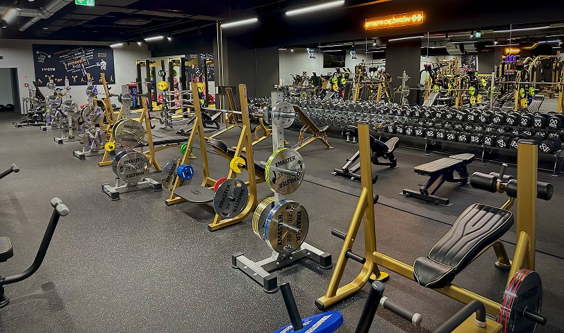 Strefa ciężarów wyposażona w ławki do wyciskania, sztangi, hantle i talerze, przeznaczona do treningu siłowego.