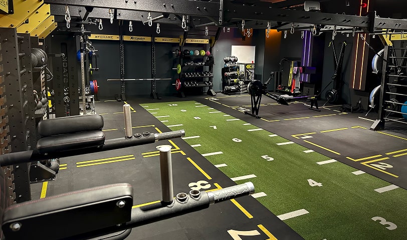 Strefa funkcjonalna z zieloną sztuczną trawą, sankami, ciężarami, sprzętem fitness i stanowiskami do różnorodnych ćwiczeń.