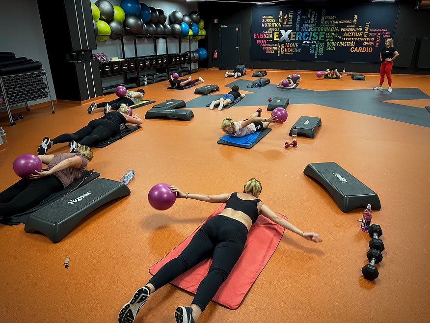 Grupowy trening fitness z instruktorem na sali – kobiety wykonują ćwiczenia w rytm muzyki.