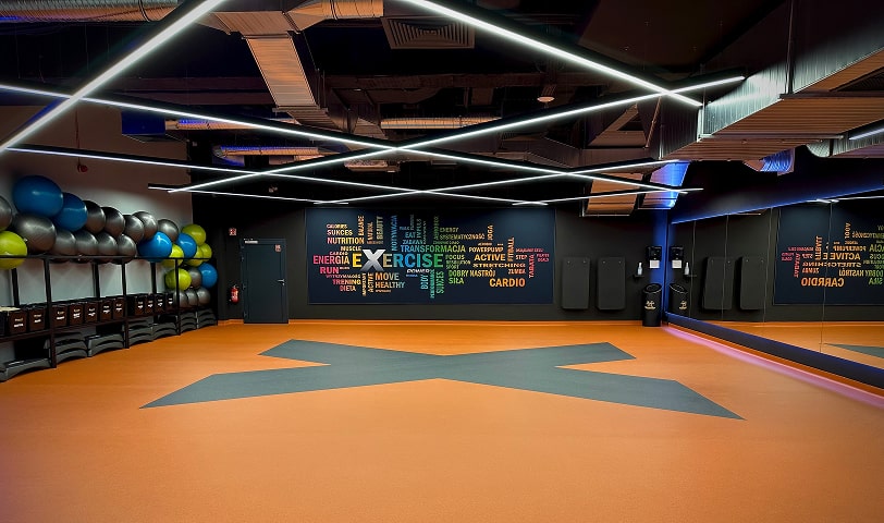 Widok na salę do zajęć fitness. Przestronne wnętrze z dużymi lustrami, sprzętem do ćwiczeń grupowych i nowoczesnym wyglądem.
